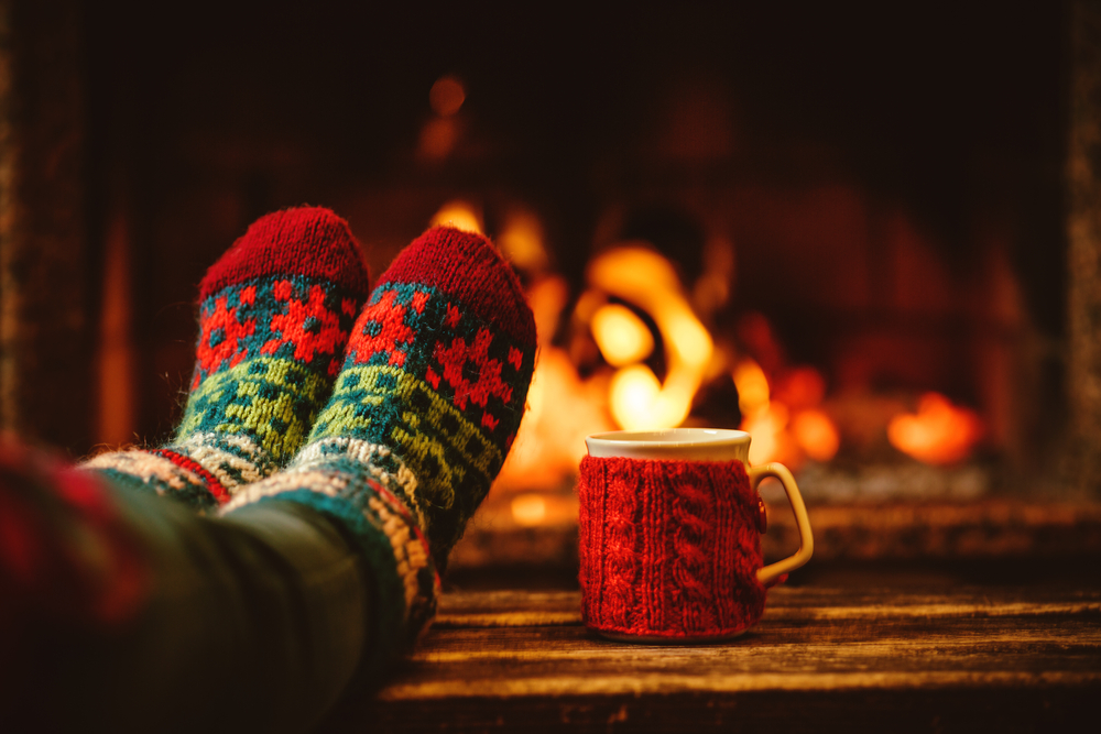 A person sits in front of the fireplace wearing snuggly holiday socks and drinking coffee/cocoa